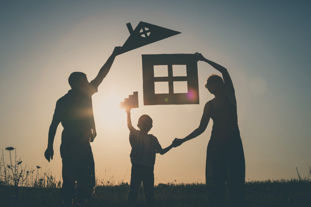日没時に屋外で家の形をした段ボールの切り抜きで遊ぶ家族のシルエットの画像。