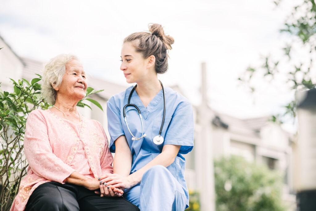 屋外の公園で、幸せな年配のアジア人女性の手を握る笑顔の看護師または介護者。