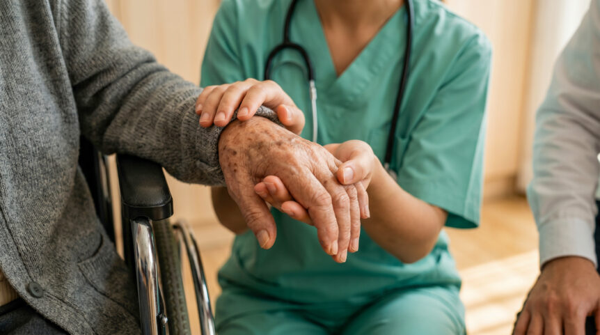 An older adult in a wheelchair has his hand held by a nurse.